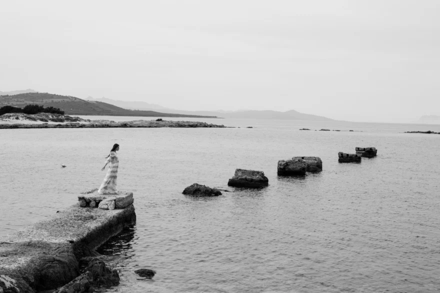 Black and white wide shot of a distant figure on rocks at the water's edge, ETRO × SHOT Magazine editorial photographed by Ina Cenusa