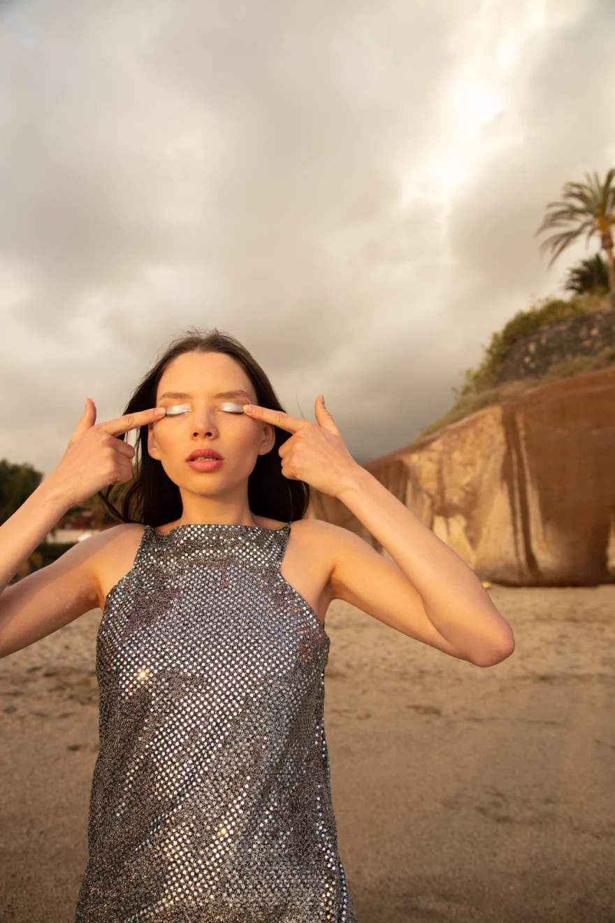 Model in a silver sequinned dress with fingers raised to her eyes, palm trees behind, Sabina in Tenerife personal project photographed by Ina Cenusa