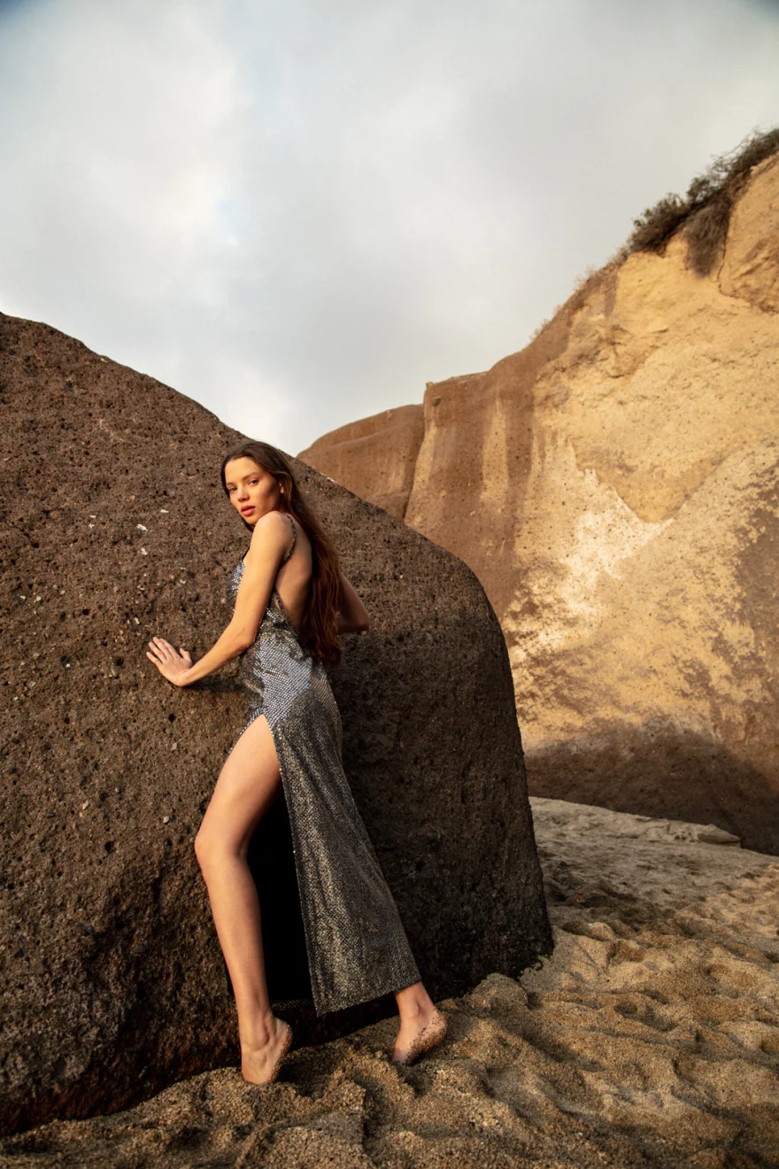 Model in a silver dress leaning against a volcanic rock on the beach, Sabina in Tenerife personal project photographed by Ina Cenusa