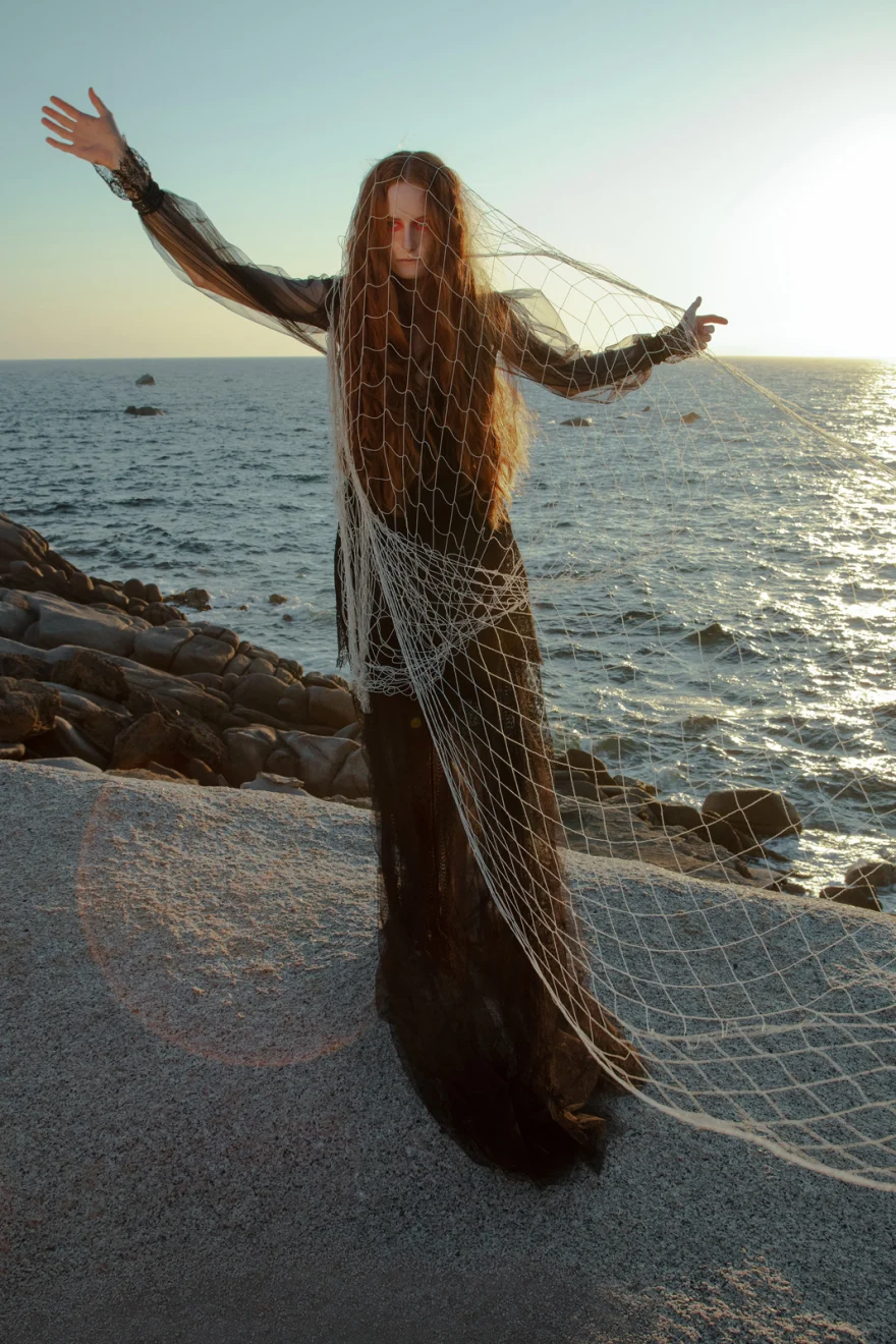 Model holding a fishing net with arms outstretched by the sea at sunset, SHOT Magazine editorial photographed by Ina Cenusa