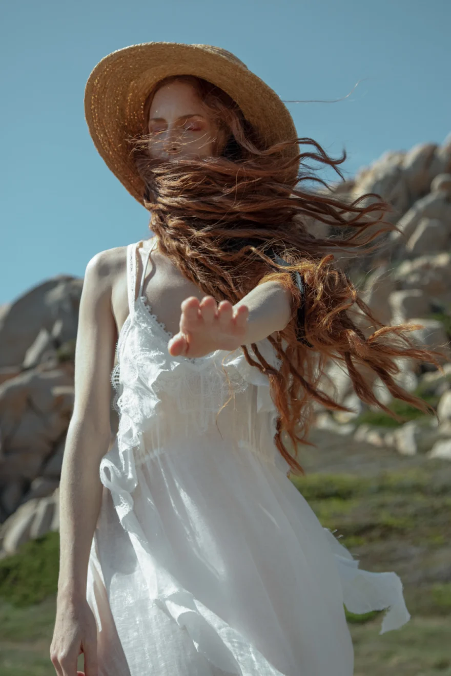 Red-haired model in a straw hat and white dress with hair blowing in the wind, SHOT Magazine editorial photographed by Ina Cenusa