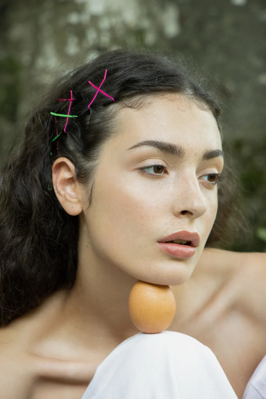 Natural-light portrait with coloured cross hair clips and an egg resting on the shoulder, SHOT Magazine editorial photographed by Ina Cenusa