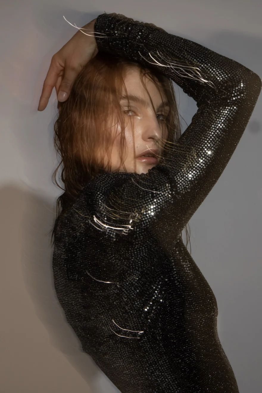 Model with arms raised in a dark sequinned dress against a pale studio wall, Iso Model Management editorial photographed by Ina Cenusa