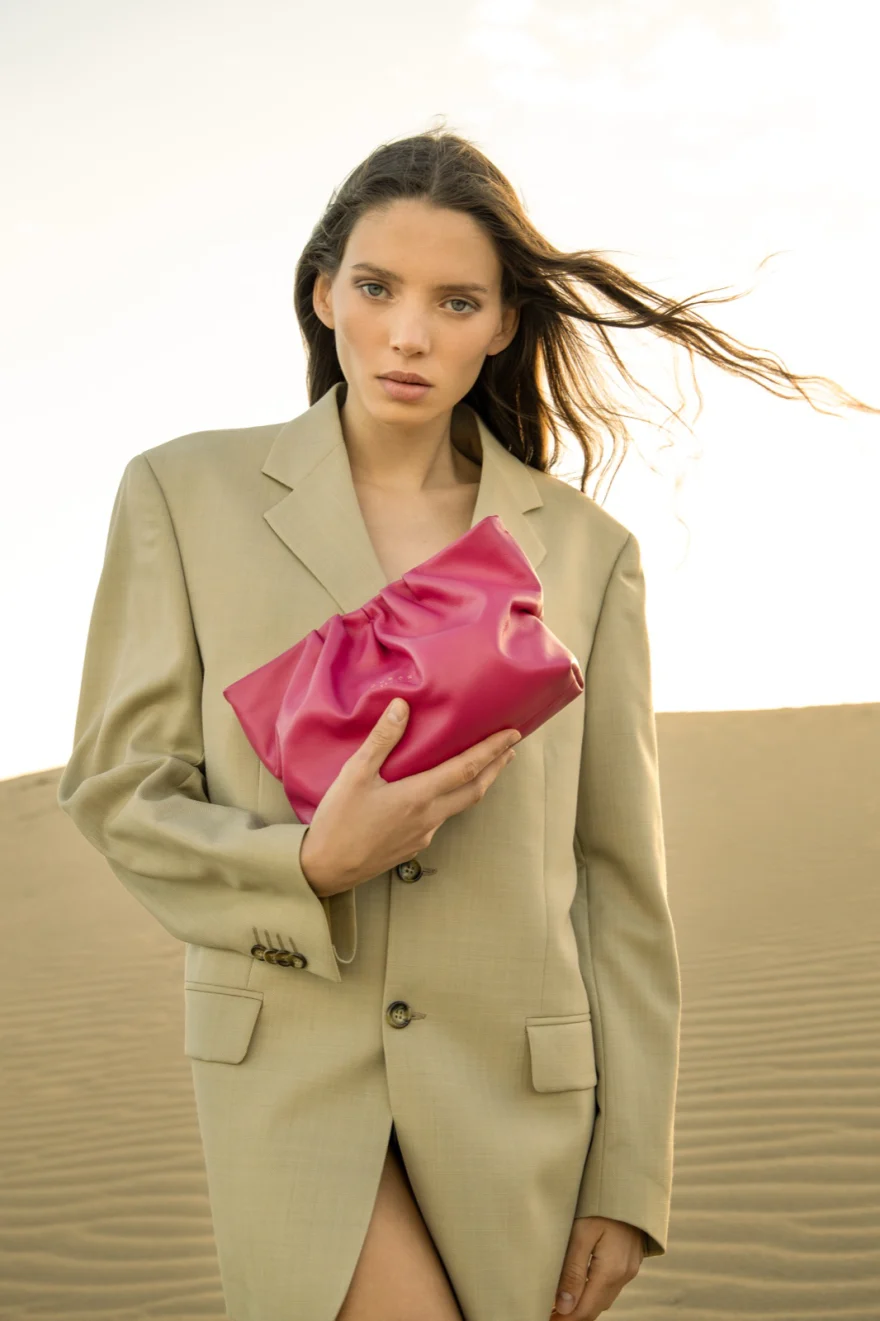 Model in a beige blazer holding a fuchsia leather clutch on the dunes, Aurora Venezia campaign photographed by Ina Cenusa