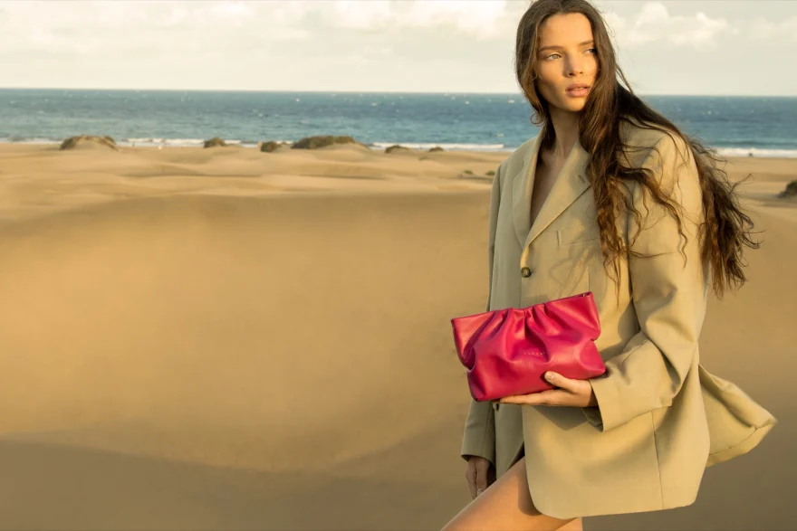Model walking the dunes with a fuchsia clutch, sea on the horizon, Aurora Venezia campaign photographed by Ina Cenusa