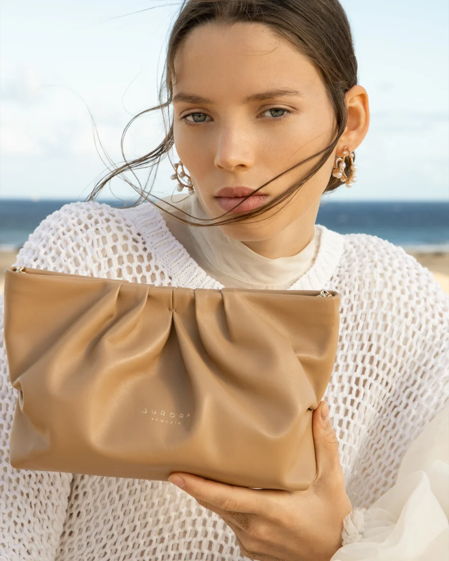 Model holding a tan leather clutch by the sea, Aurora Venezia campaign photographed by Ina Cenusa