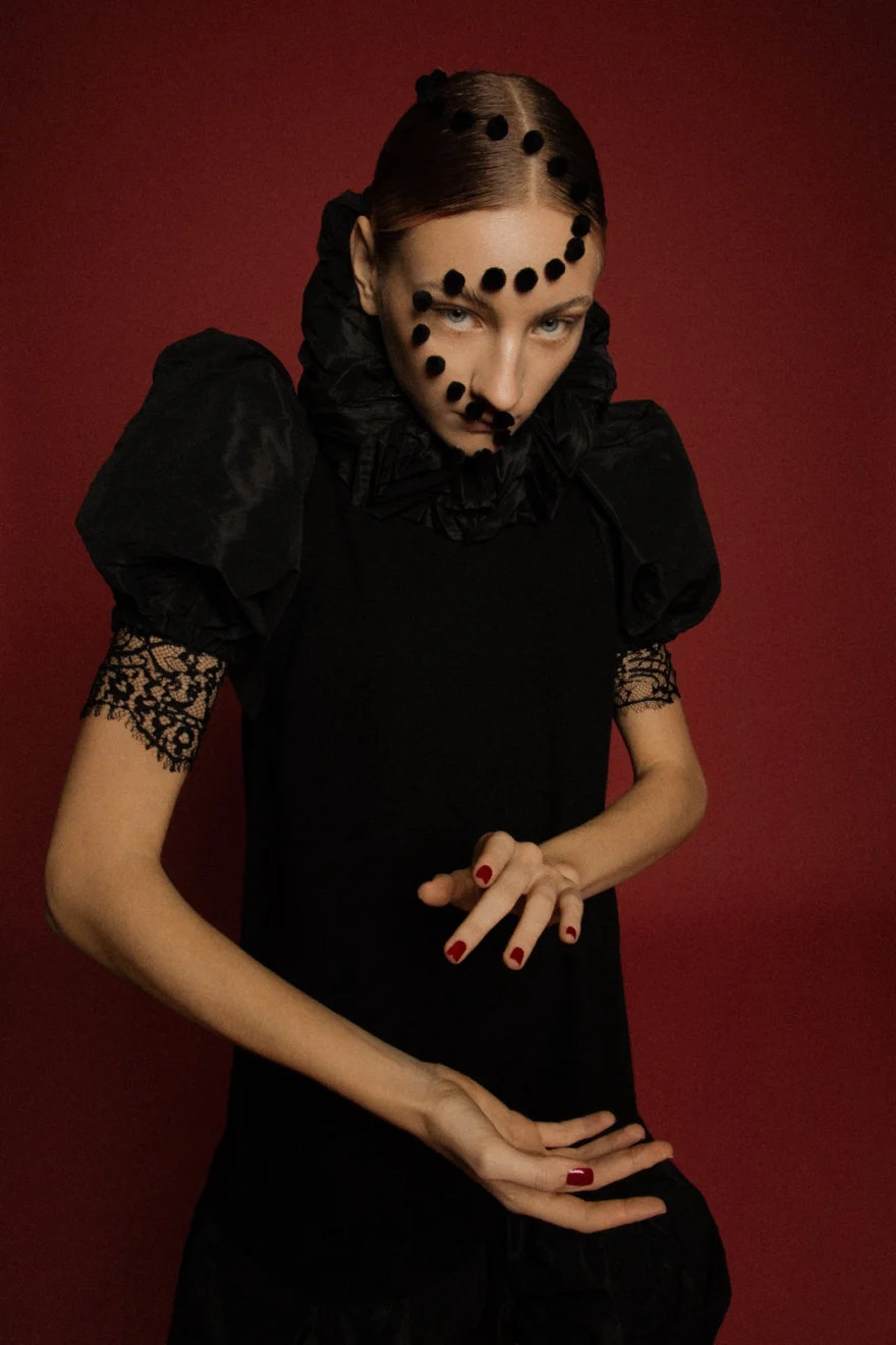 Model in a black dress and pom-pom veil with hands raised, red backdrop, LYX Magazine editorial photographed by Ina Cenusa