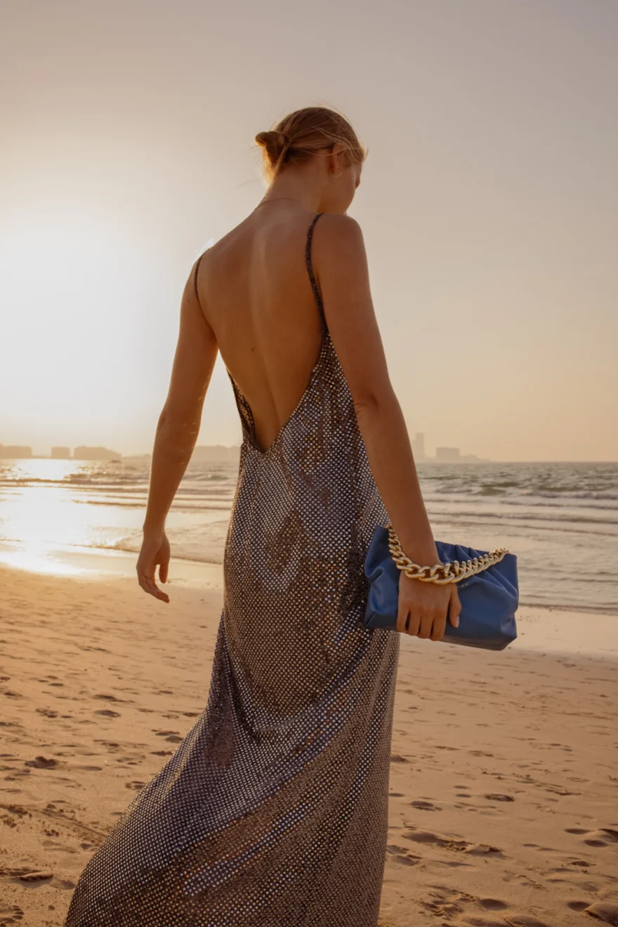Model seen from behind in a backless sequin dress carrying a blue chain bag along the shore, Aurora Venezia campaign in Dubai photographed by Ina Cenusa
