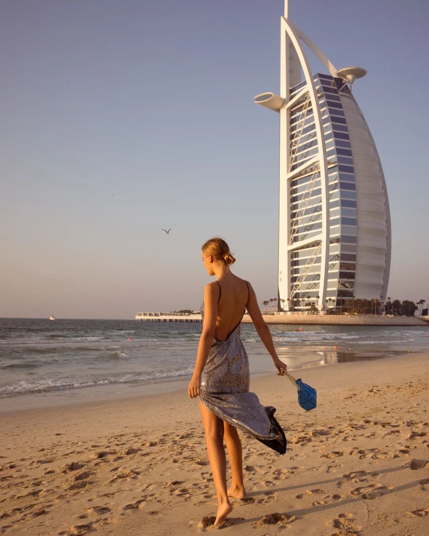 Model walking the beach with a blue bag and the Burj Al Arab behind her at sunset, Aurora Venezia campaign in Dubai photographed by Ina Cenusa