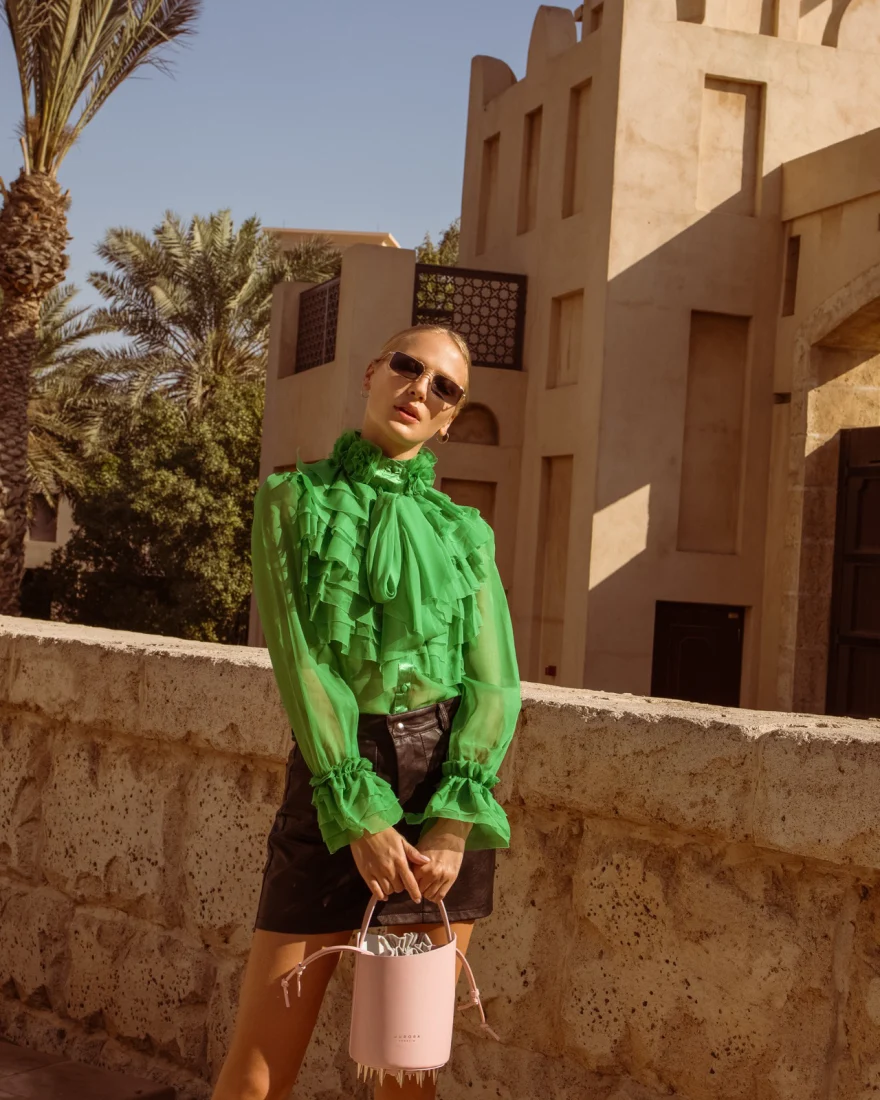 Model in a green ruffled blouse and sunglasses holding a pink bucket bag in a desert village, Aurora Venezia campaign in Dubai photographed by Ina Cenusa