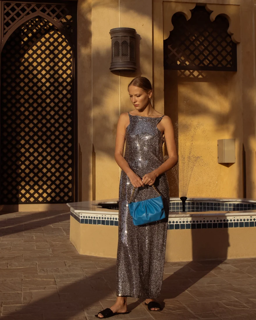 Model in a silver sequin dress holding a blue quilted bag in front of carved mashrabiya screens, Aurora Venezia campaign in Dubai photographed by Ina Cenusa