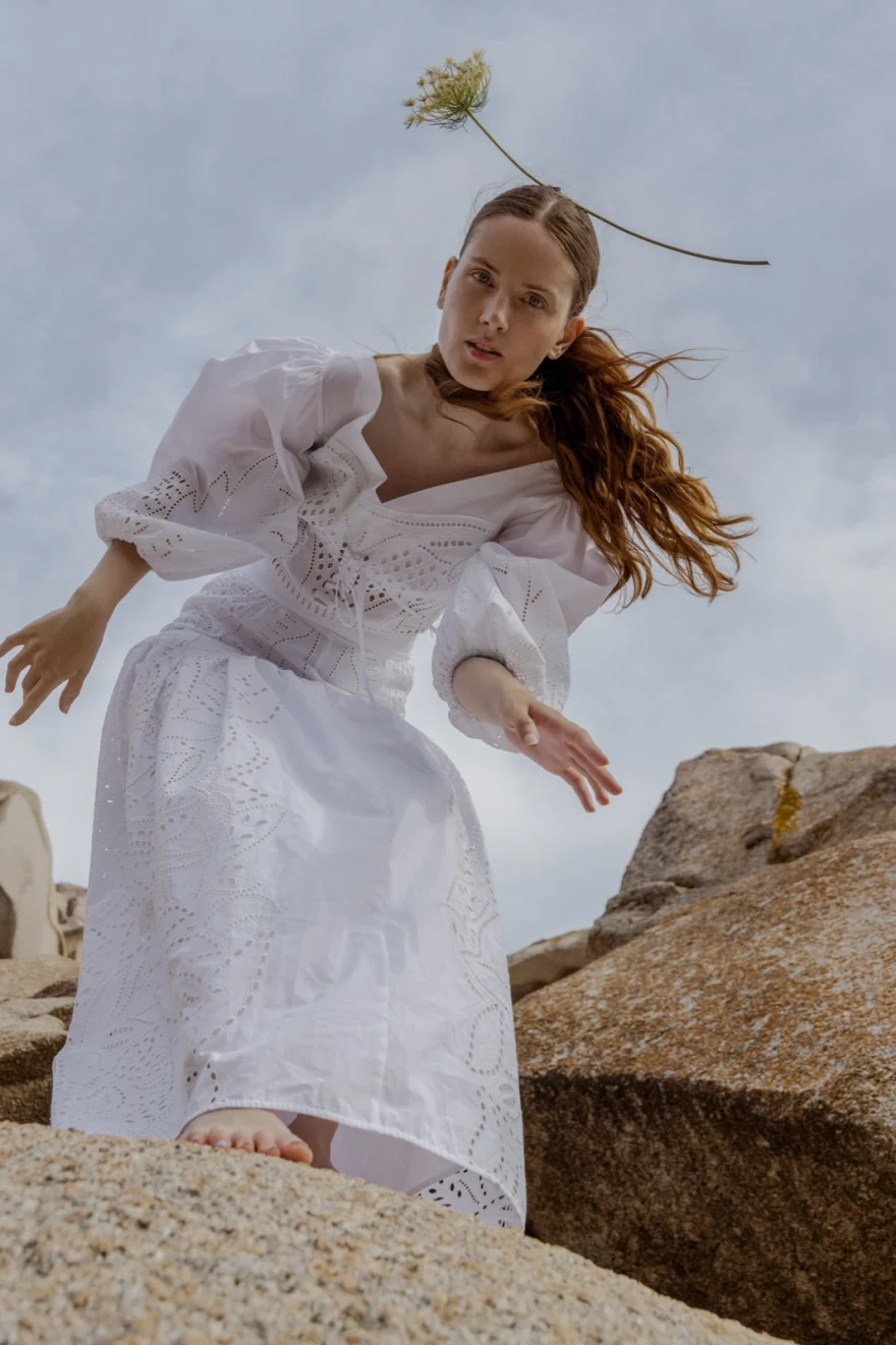 Model in a white broderie dress stepping across granite rocks with a flower in her hair, SHOT Magazine editorial photographed by Ina Cenusa