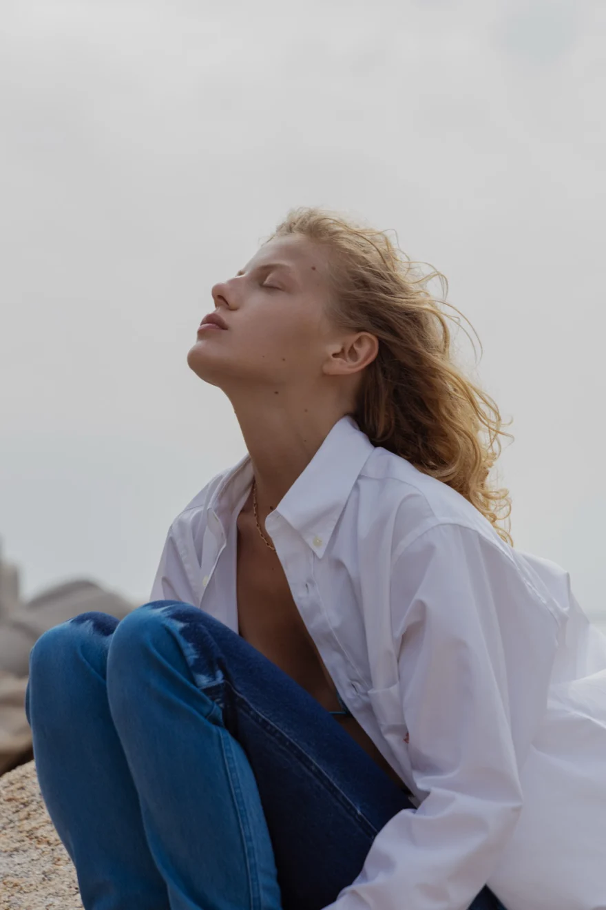 Model in an open white shirt and denim seated on the rocks against a pale sky, SHOT Magazine editorial photographed by Ina Cenusa