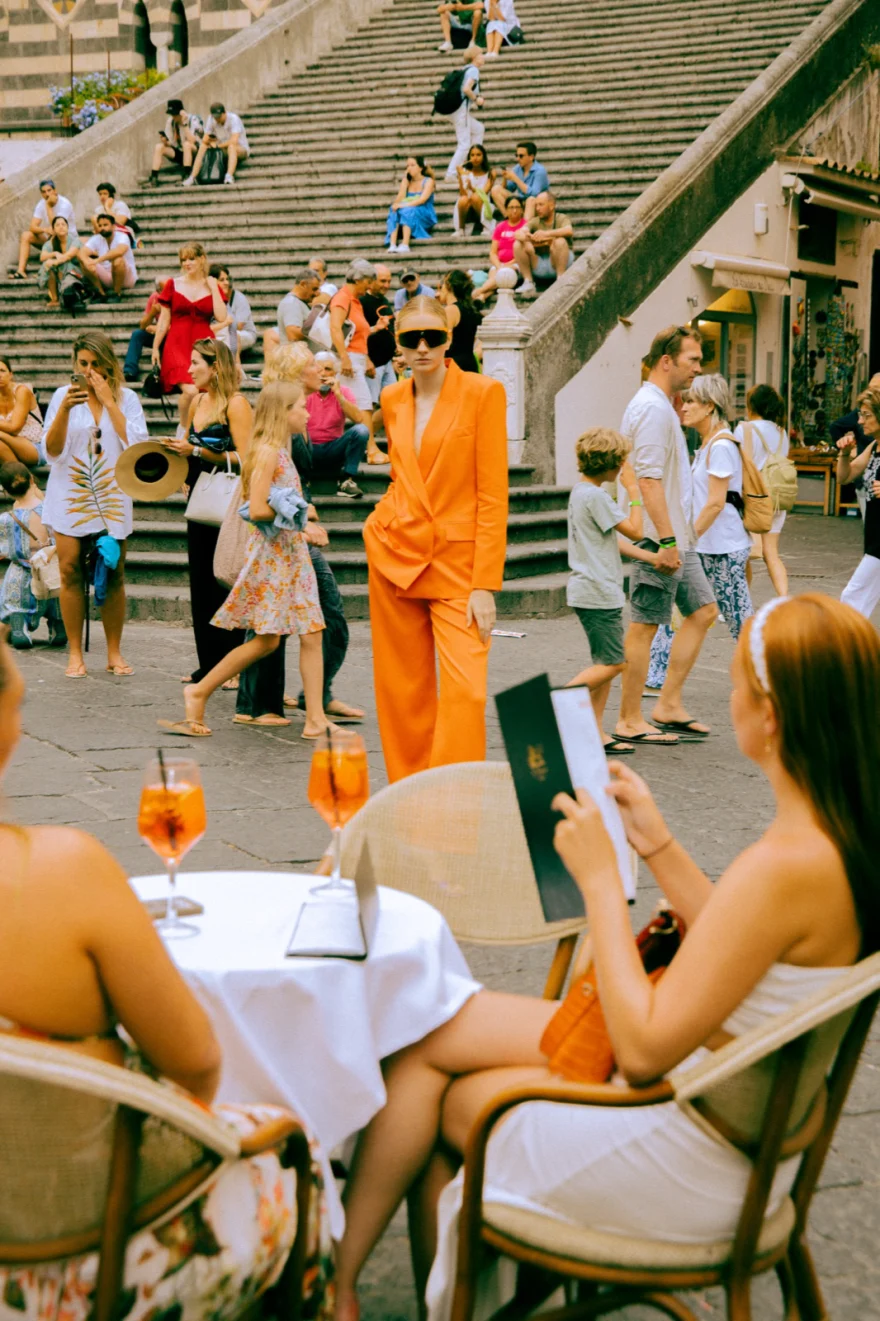 Model in an orange suit seen past café tables in the square of Amalfi, Amalfi Coast personal project photographed by Ina Cenusa