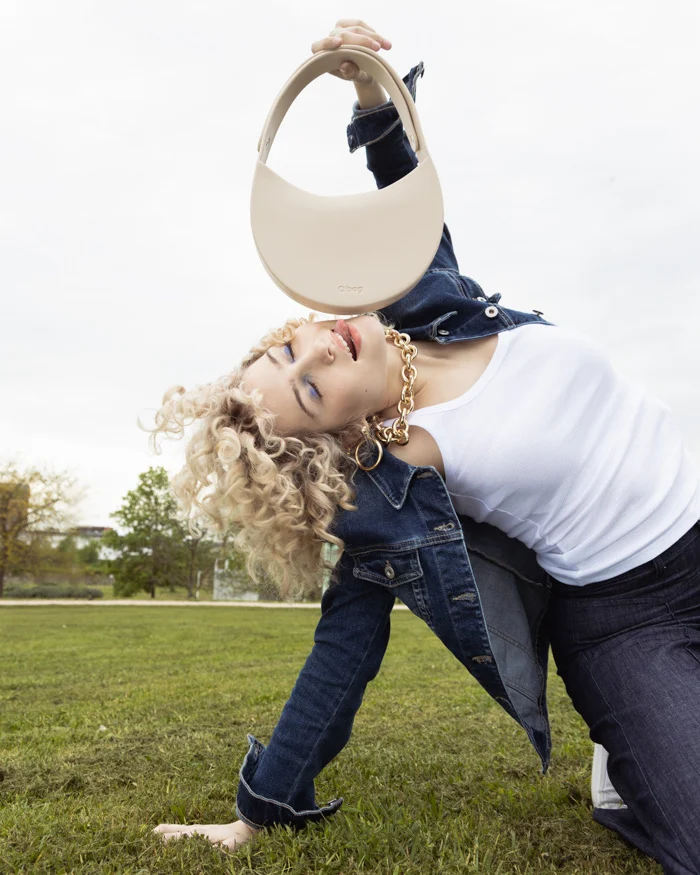 Model with curly blonde hair bending backwards holding a cream bag, Carrera Jeans campaign photographed by Ina Cenusa