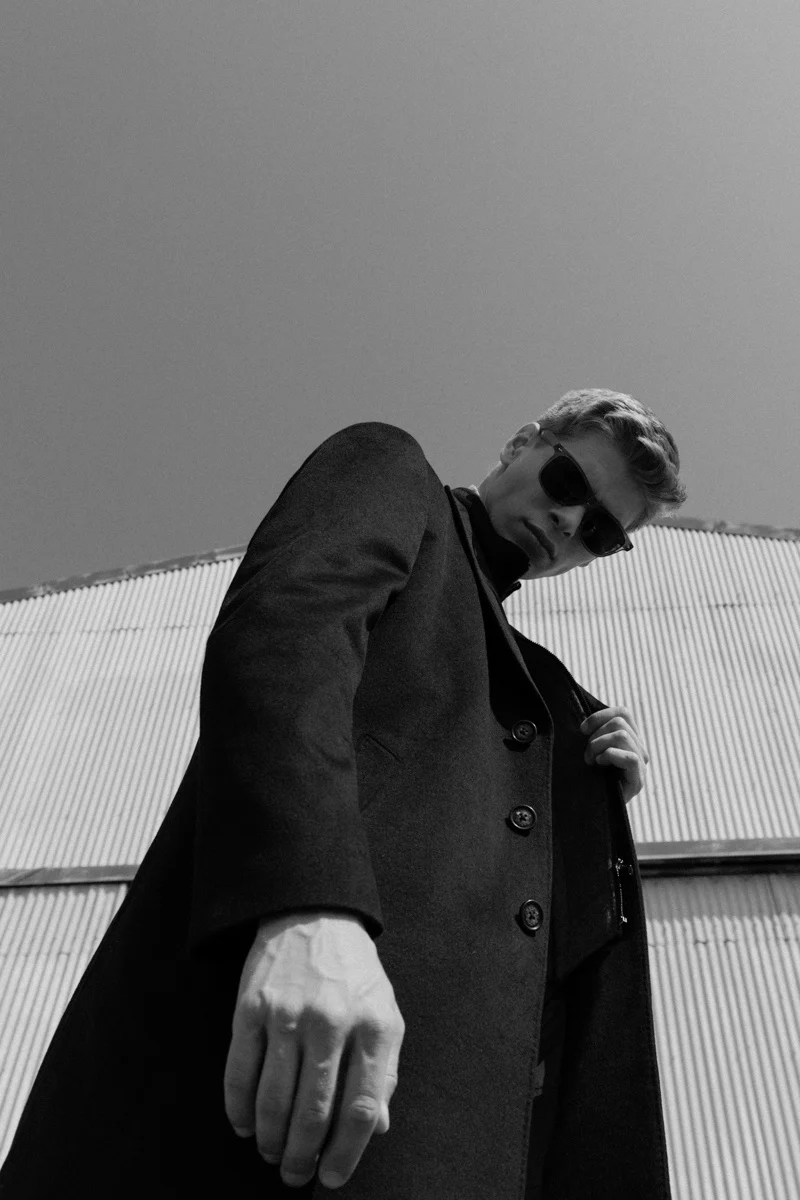 Man in a long dark coat leaning against a metal wall, Mabrun campaign photographed by Ina Cenusa