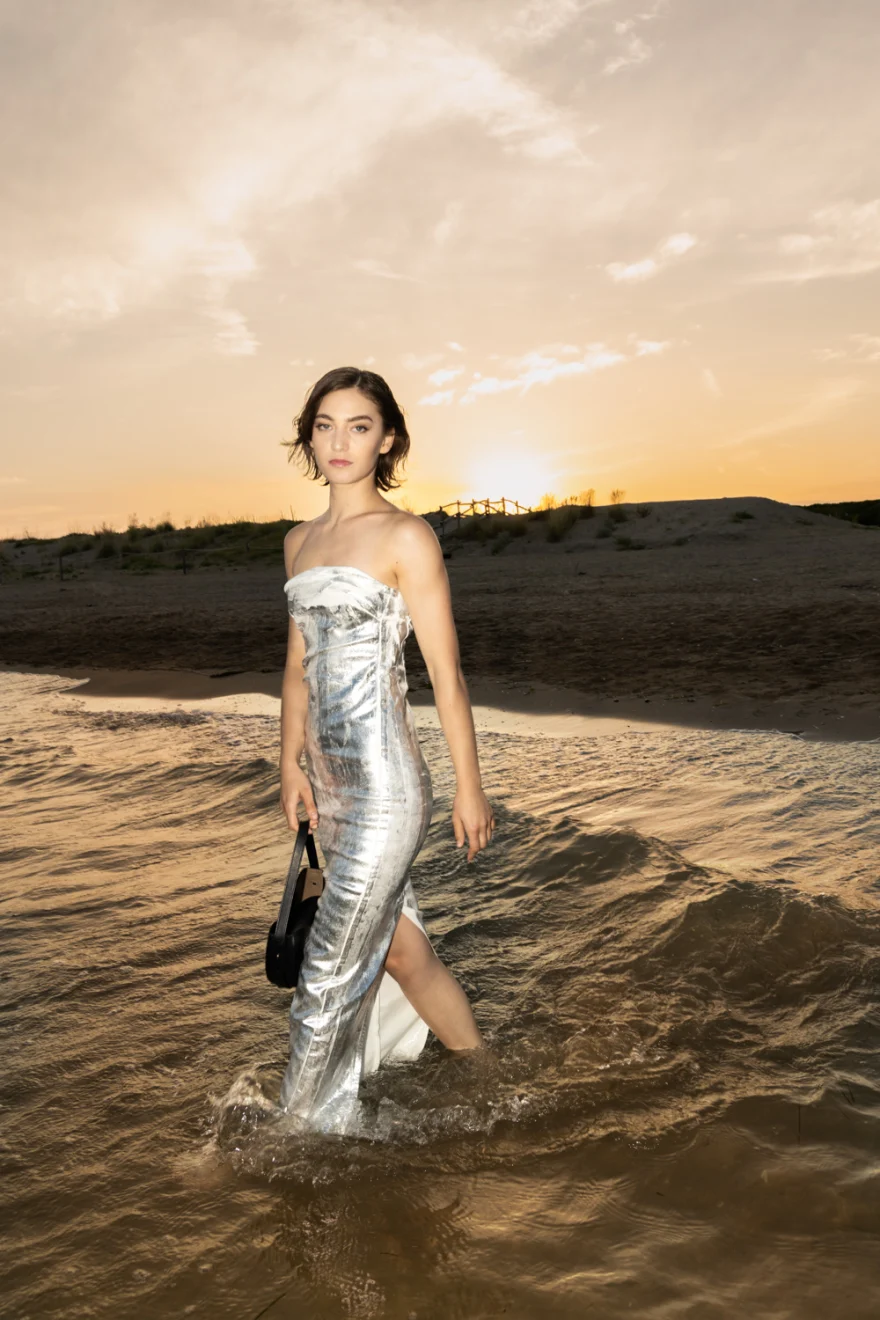 Model in a silver dress wading at sunset with a black bag, Noesi campaign photographed by Ina Cenusa