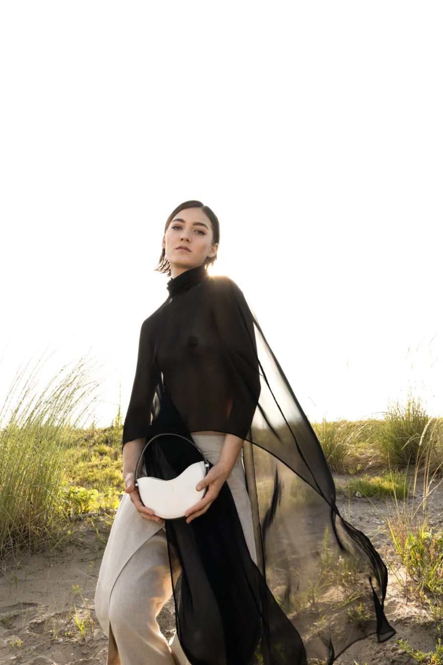 Model in sheer black on the dunes with a white crescent bag, Noesi campaign photographed by Ina Cenusa