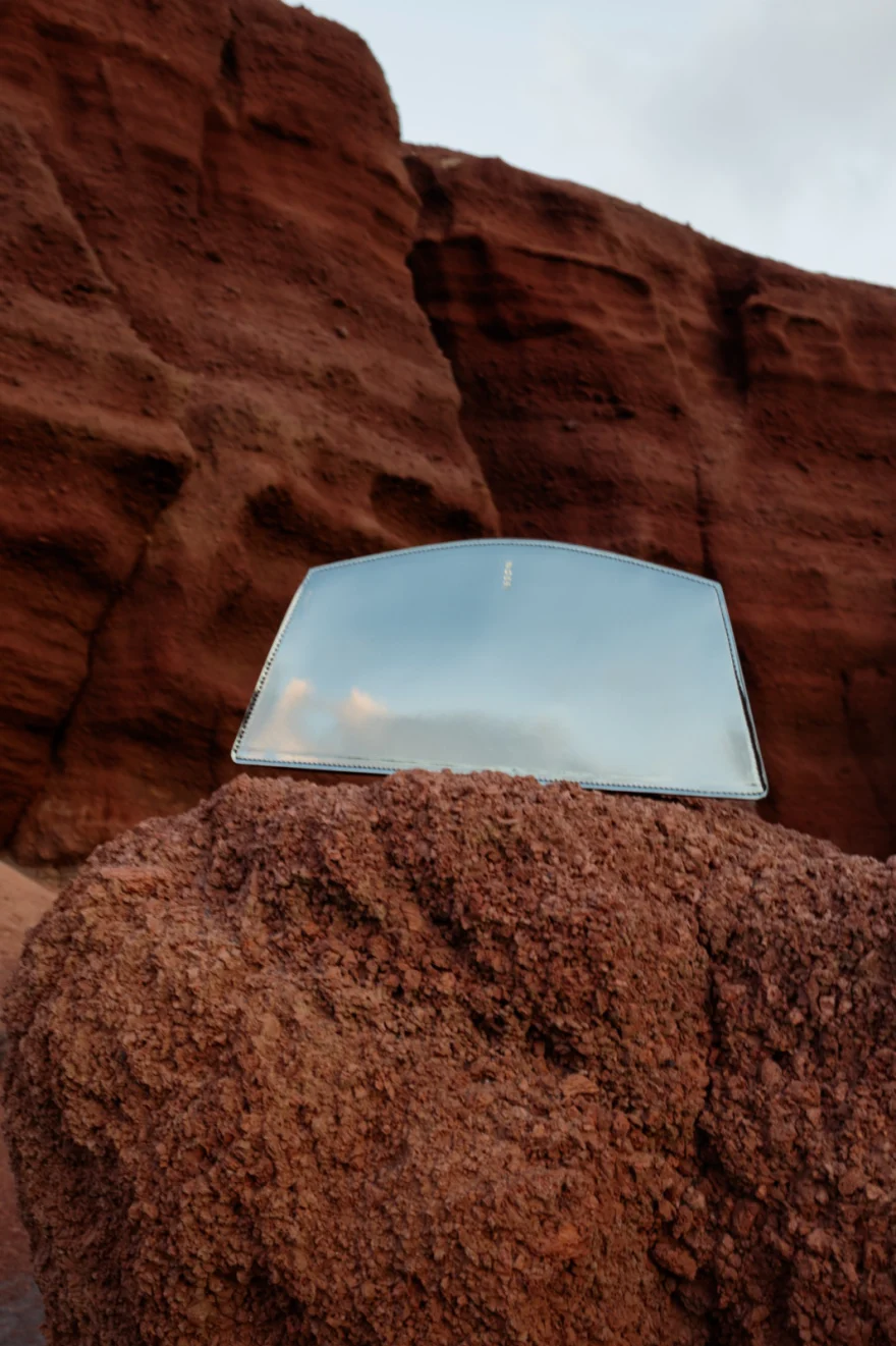 Mirror finish clutch reflecting the sky, resting on red volcanic rock, Noesi campaign photographed by Ina Cenusa