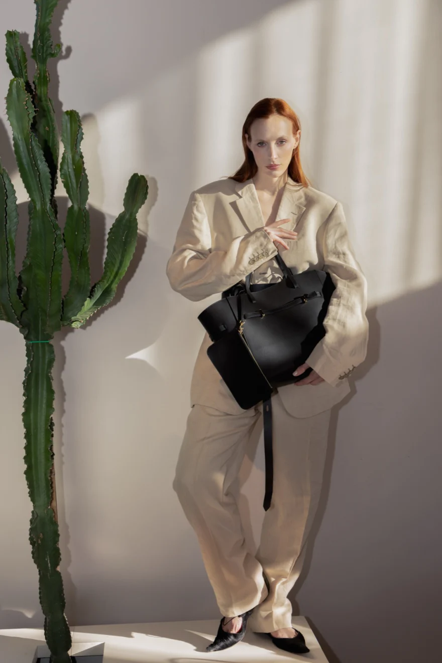 Model in a cream coat holding a black leather bag beside a plant, Biebi campaign photographed by Ina Cenusa