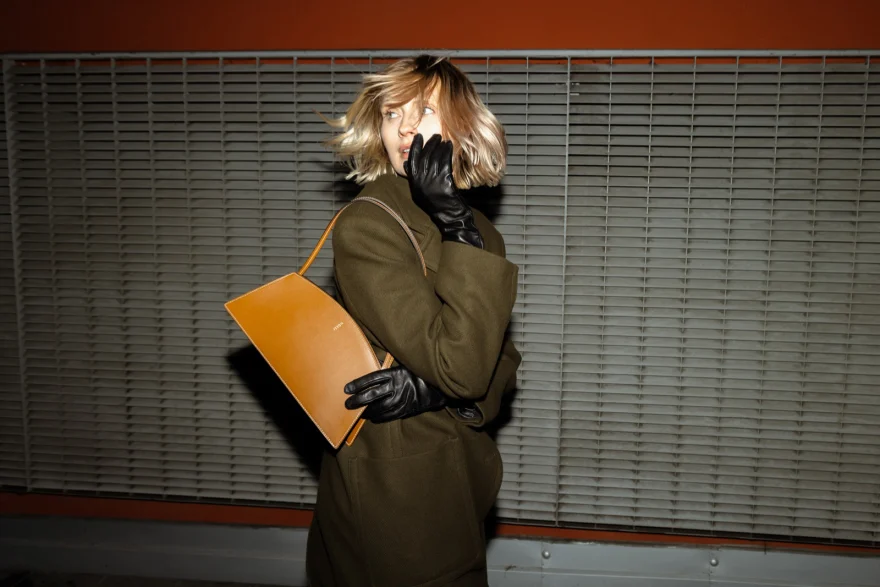 Model in an olive coat holding a tan clutch against a shutter, Noesi campaign photographed by Ina Cenusa