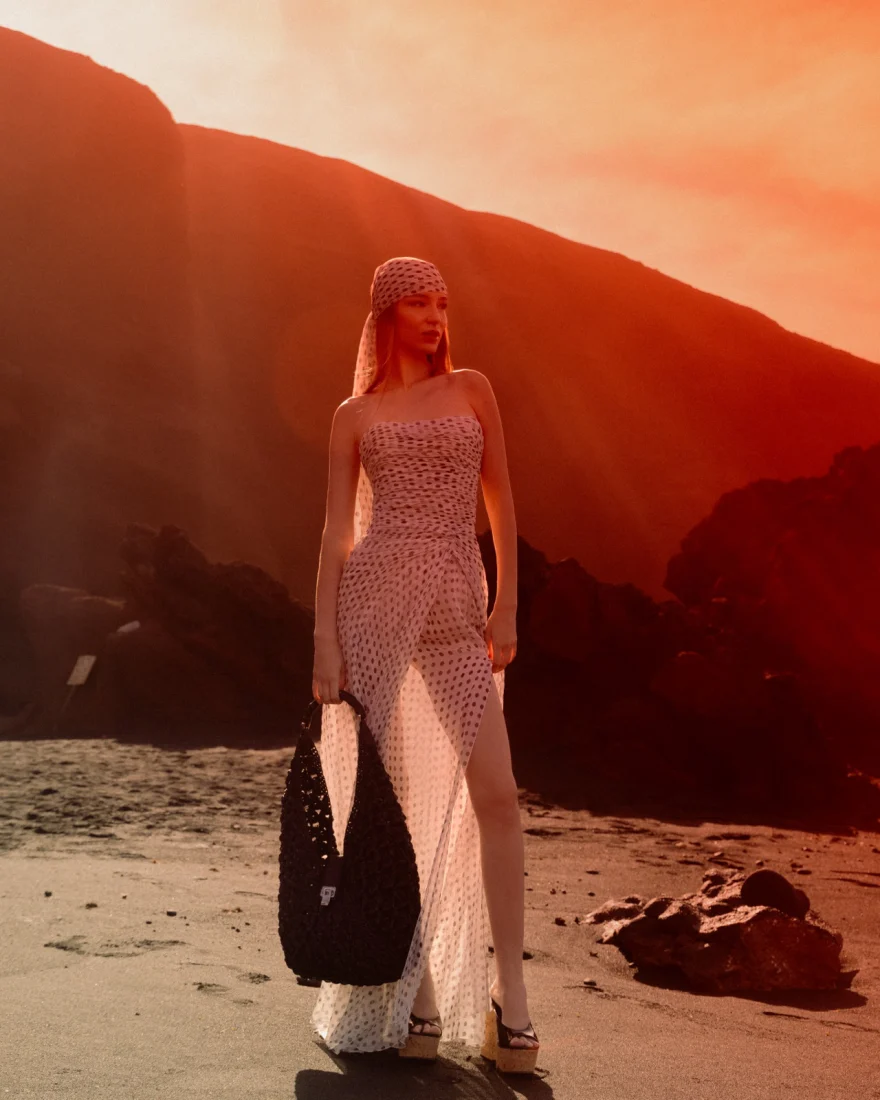Model in a sheer polka-dot dress and headscarf holding a black crochet bag on a volcanic beach at sunset, Ermanno Scervino campaign photographed by Ina Cenusa