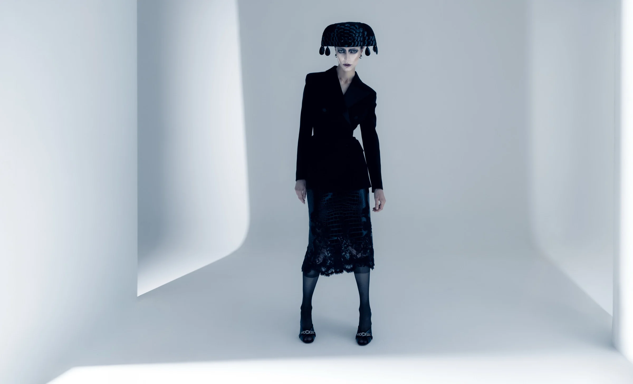 Model in black tailoring and a sculpted headpiece in a white studio corner, editorial by fashion photographer Ina Cenusa for SHOT Magazine