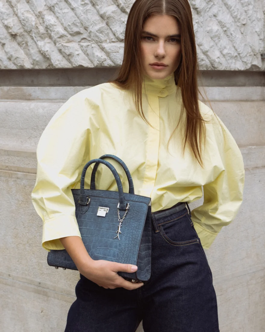 Model in a pale yellow shirt holding a blue crocodile effect tote, Pagani campaign in Geneva photographed by Ina Cenusa